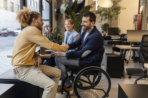Businessman in wheelchair doing handshake with colleague at co-working space - JCCMF11289