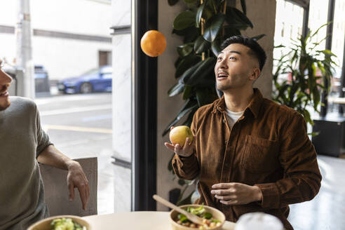 Geschäftsmann spielt mit Apfel und Orange im Bürocafé sitzend - JCCMF11315