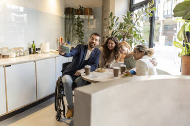 Businessman in wheelchair taking selfie with colleagues sitting at office cafe - JCCMF11317