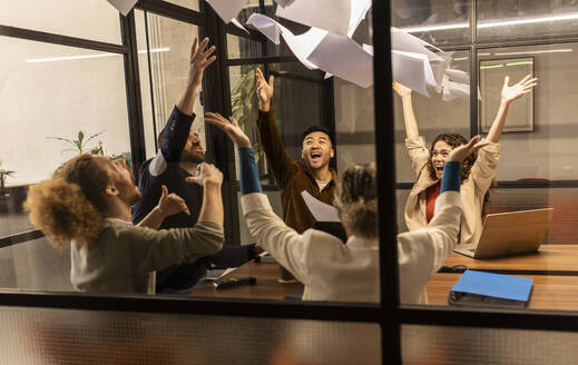 Cheerful business colleagues throwing papers seen through glass window at office - JCCMF11326