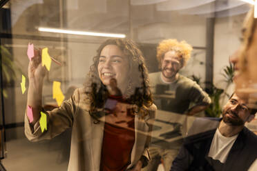 Smiling businesswoman doing brainstorming session with colleagues behind glass at office - JCCMF11328