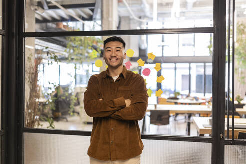 Smiling young businessman standing with arms crossed at office - JCCMF11345