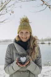 Happy woman holding heart shaped snowball near lake - KVBF00004