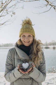 Glückliche Frau hält herzförmigen Schneeball in der Nähe eines Sees - KVBF00004