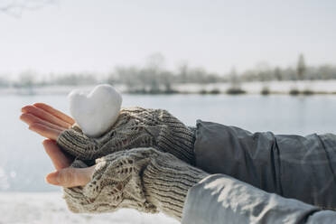 Hands of woman holding heart shaped snowball near lake - KVBF00005