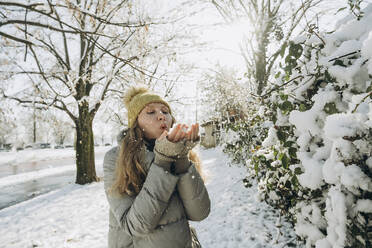 Verspielte Frau bläst mit der Hand auf Schnee in der Nähe von Pflanzen - KVBF00008