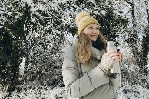 Lächelnde Frau hält Flasche in der Nähe von Bäumen im Schnee bedeckt - KVBF00009