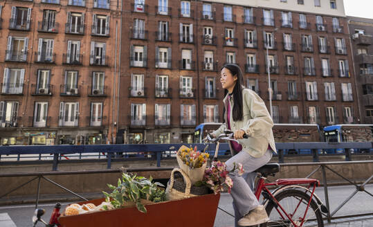 Smiling young woman riding cargo bike near building in city - JCCMF11402