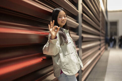 Smiling woman gesturing OK sign and leaning on shutter - JCCMF11406