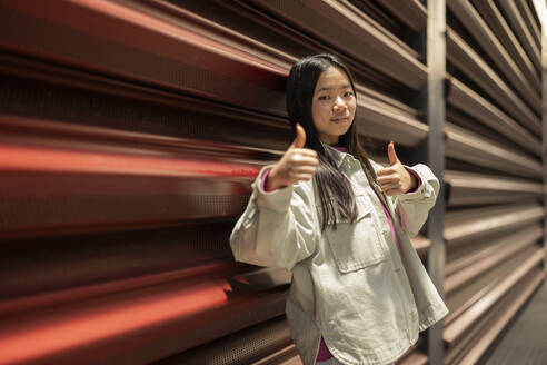 Young woman showing thumbs up gesture and leaning on shutter - JCCMF11407