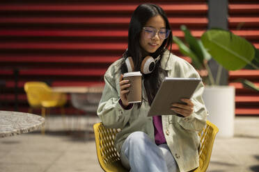 Smiling young woman holding coffee cup and using tablet PC at sidewalk cafe - JCCMF11411