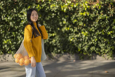 Smiling woman standing with bag of oranges near plants - JCCMF11416