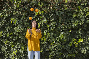 Glückliche junge Frau jongliert mit Orangen vor einer Pflanze - JCCMF11418