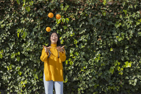 Happy young woman juggling oranges in front of plants - JCCMF11418