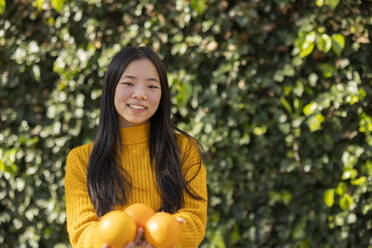 Lächelnde junge Frau mit Orangen in der Hand vor einer Pflanze - JCCMF11419