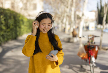 Lächelnde Frau, die drahtlose Kopfhörer trägt und ein Smartphone benutzt - JCCMF11422