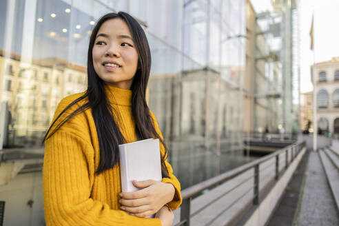 Happy woman standing with book near glass building - JCCMF11433