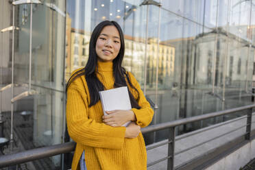 Smiling woman standing with book near glass building - JCCMF11434