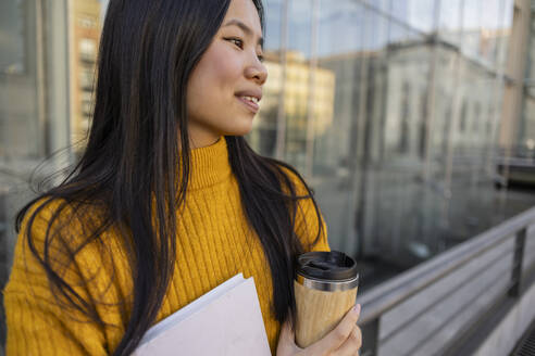 Lächelnde junge Frau steht mit Buch und Kaffeetasse in der Nähe eines Gebäudes - JCCMF11436
