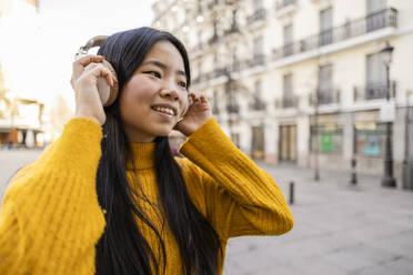 Happy young woman wearing wireless headphones near building - JCCMF11439