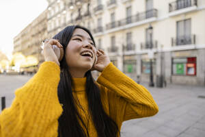 Fröhliche junge Frau mit drahtlosen Kopfhörern in der Nähe eines Gebäudes - JCCMF11440