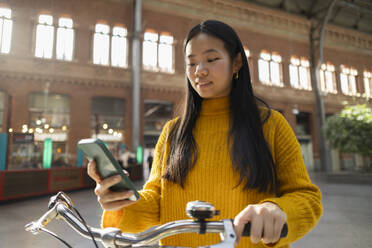 Smiling woman with bicycle using smart phone at station - JCCMF11450