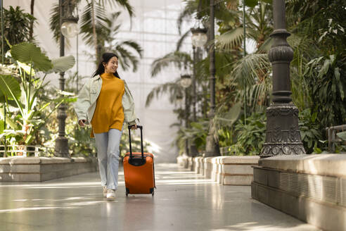 Glückliche junge Frau geht mit Koffer in der Nähe von Pflanzen - JCCMF11460