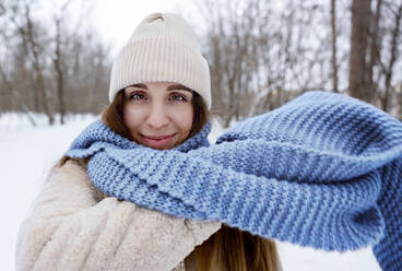 Smiling woman wearing knitted hat and blue scarf in winter forest - MBLF00271