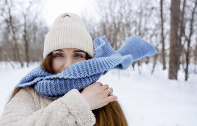 Young woman wearing knit hat and blue scarf in winter forest - MBLF00272