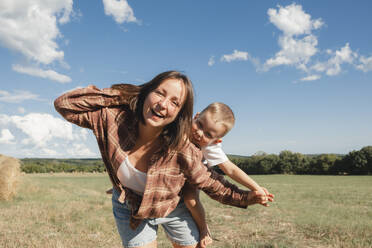 Verspielte Frau, die ihren Sohn auf einem Feld an einem sonnigen Tag huckepack nimmt - KVBF00016
