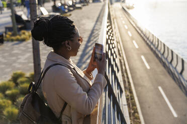 Woman taking picture of river through smart phone at sunset - IKF01726