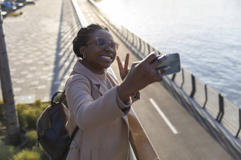 Lächelnde Frau gestikuliert Friedenszeichen und nimmt Selfie am Fluss - IKF01727