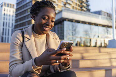 Smiling woman sitting on stairs and using smart phone in city at sunset - IKF01748