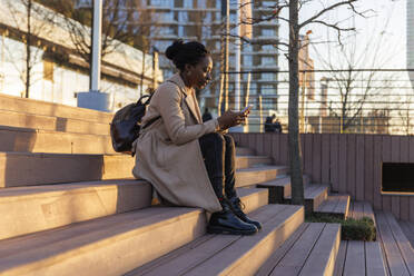 Woman using smart phone on stairs in city at sunset - IKF01751