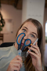 Playful young businesswoman holding scissors over face and talking on smart phone in office - JOSEF23479