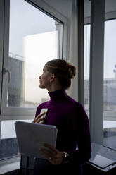Businesswoman holding documents and looking out of window in office - JOSEF23506