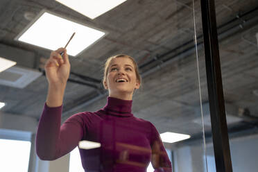 Glückliche Geschäftsfrau hält Stift in der Nähe von Glaswand im Büro - JOSEF23515