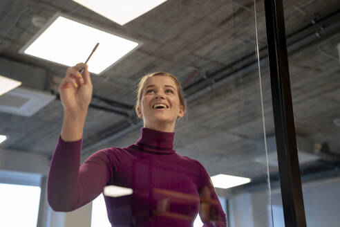 Happy businesswoman holding pen near glass wall in office - JOSEF23515