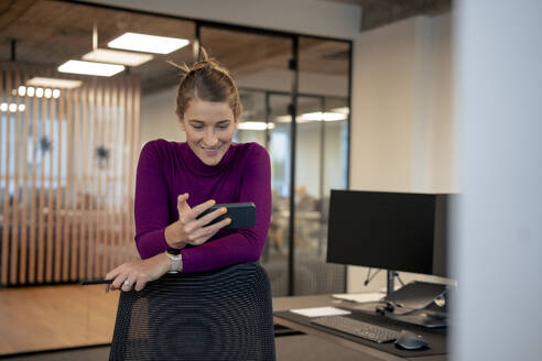 Smiling businesswoman using smart phone and leaning on chair in office - JOSEF23517