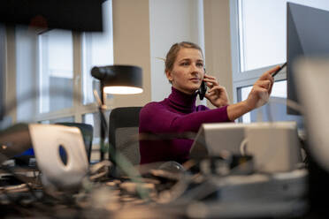 Geschäftsfrau spricht mit einem Smartphone und zeigt mit einem Stift auf einen Computer im Büro - JOSEF23522