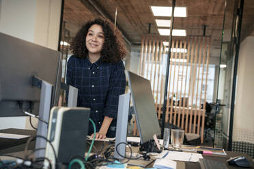 Lächelnde Geschäftsfrau lehnt sich an einen Schreibtisch in der Nähe von Computern im Büro - JOSEF23536
