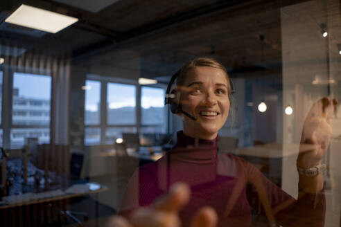 Happy customer service representative wearing headset near glass wall in office - JOSEF23546