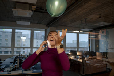 Playful businesswoman talking on smart phone and catching balloon in office - JOSEF23554