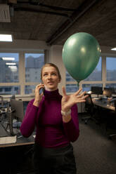 Businesswoman talking on smart phone and catching balloon in office - JOSEF23558