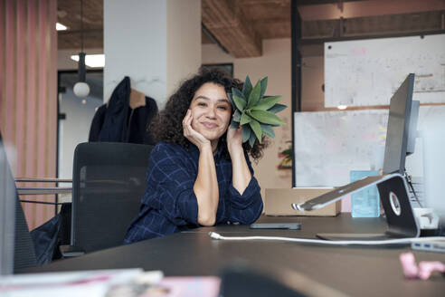Smiling businesswoman holding plant near face at desk in office - JOSEF23563