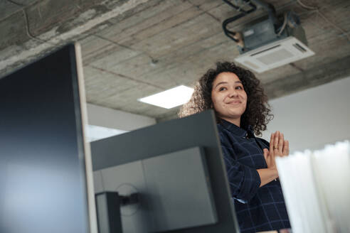 Lächelnde Geschäftsfrau, stehend mit gefalteten Händen in der Nähe eines Computers im Büro - JOSEF23566