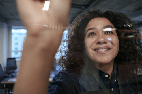 Happy businesswoman with curly hair near transparent glass in office - JOSEF23573