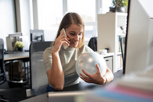 Lächelnde Geschäftsfrau, die einen Ballon hält und mit einem Smartphone im Büro telefoniert - JOSEF23589