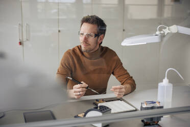 Thoughtful engineer working with soldering iron at workshop - JOSEF23615