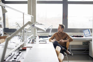 Thoughtful mature engineer sitting on chair near desk at workshop - JOSEF23620